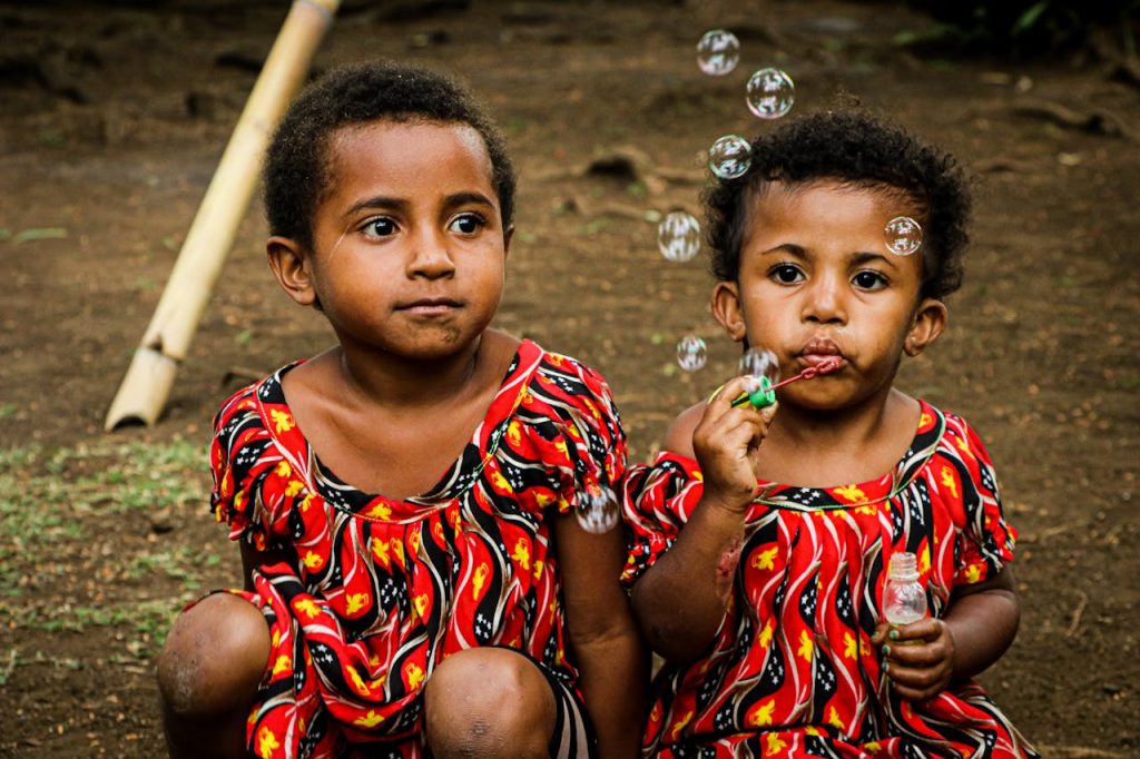 fashion-man-person-couple-10404339 Two young children in vibrant traditional attire blowing bubbles outdoors in Port Moresby.