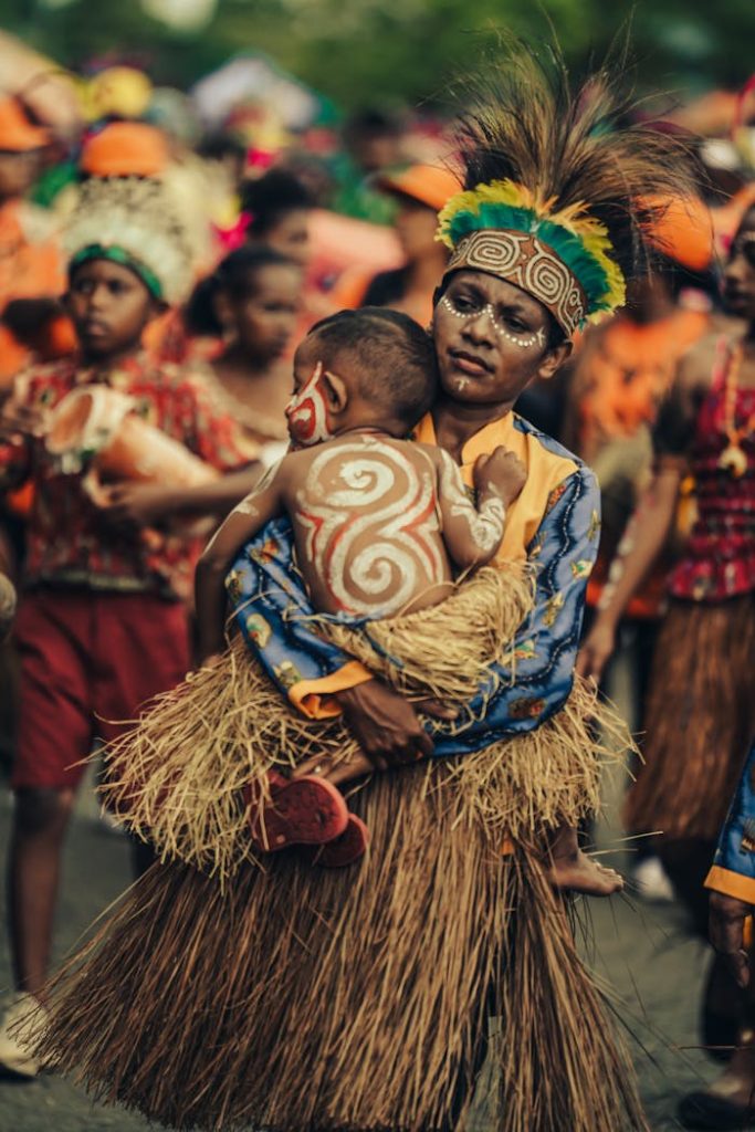 parent-holding-child-on-traditional-parade-13454788 Celebrating Papuan tradition with colorful attire and body art in a community parade.